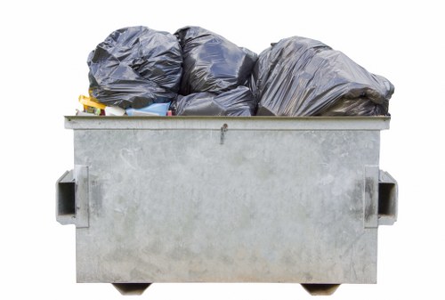 A large, rectangular metal rubbish skip positioned outdoors on a paved surface, filled with multiple black garbage bags made from plastic, which are visibly tied at the tops. The bags are arranged haphazardly, with some leaning on each other, and occupy most of the skip's interior space. The metallic surface of the skip appears weathered, with a slightly rough texture and a dull grey finish. In the background, part of the skip's side handles is visible, and the scene suggests a typical waste collection setting in an urban or suburban area, possibly near residential or commercial properties. The overall lighting indicates a neutral daylight environment, supporting the context of rubbish removal services such as those offered by Bayswater House Clearance, which operates in the London area including Bayswater postcode regions.