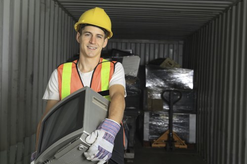 A young male waste management worker, wearing a yellow safety helmet, high-visibility orange and green vest, and white gloves with purple accents, is inside a metallic storage unit or container with corrugated steel walls. He is smiling and holding a gray, slightly curved electronic device or appliance, possibly a television or monitor, which is partially lifted or carried. In the background, there are several large, black plastic-wrapped pallets or boxes stacked against the container wall, and a yellow hydraulic pallet jack with a black handle positioned towards the back, indicating a rubbish removal or clearance operation. The environment is well-lit, likely by natural light, and the setting suggests an organized space used for waste collection or disposal in a town area. This setup aligns with commercial rubbish removal services, such as those offered by Bayswater House Clearance, operating in the Bayswater or nearby postcode region.